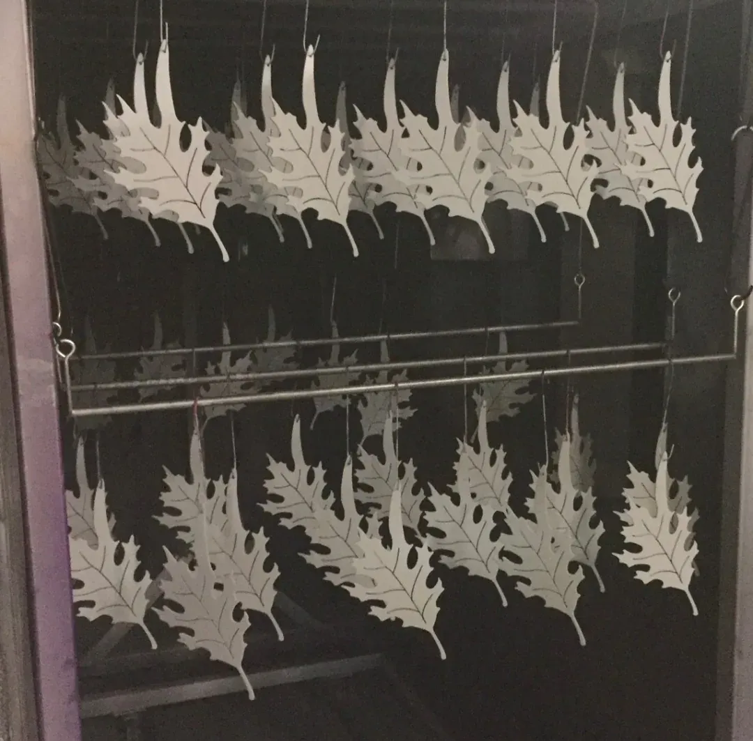Row of laser-cut steel oak leaves with the veining cut through, hanging on wires in the coating booth
